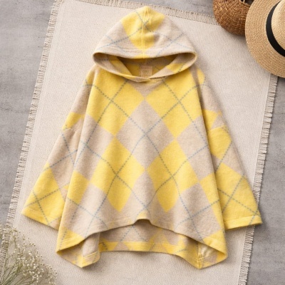 Capa amarelo e bege xadrez com capuz sobre tapete bege com franjas, chapéu e cesta ao lado.