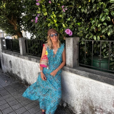 Mulher com vestido longo azul floral encostada em parede de cimento com plantas ao fundo