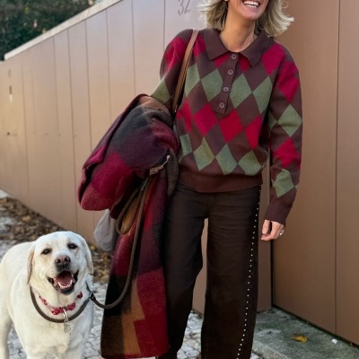 Mulher com camisola xadrez vermelha e verde, calças castanhas com tachas, casaco xadrez e óculos de sol, ao lado de cão Labrador branco.
