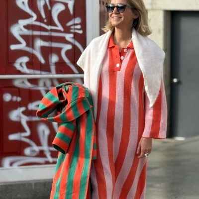 Mulher com vestido de riscas verticais cor de rosa e laranja e casaco de riscas horizontais verdes e laranja.