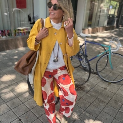 Mulher a usar casaco amarelo, calças com padrão floral, óculos de sol e bolsa castanha numa rua urbana
