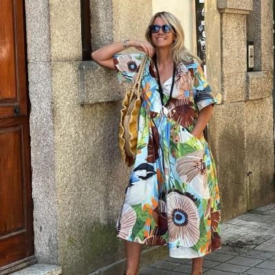 Mulher com vestido midi floral colorido, bolsa de palha e sapatos castanhos numa rua com parede de pedra