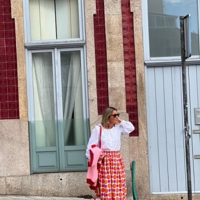 Mulher vestida com camisola branca e saia colorida em calçada urbana junto a parede de azulejos vermelhos