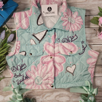 Colete acolchoado azul com flores cor-de-rosa, corações e escrita preta, com botões e laços verdes nas laterais.