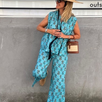 Mulher com conjunto azul floral, chapéu de palha, óculos escuros, mala castanha e sapatos de palha junto a uma parede cinzenta
