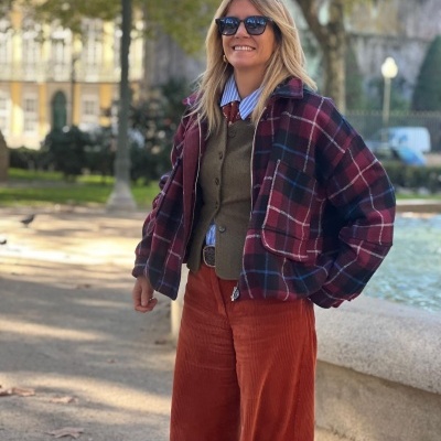 Mulher com casaco xadrez bordeaux, azul e branco, camisola azul às riscas, colete verde-oliva e calças largas laranja de veludo cotelê.
