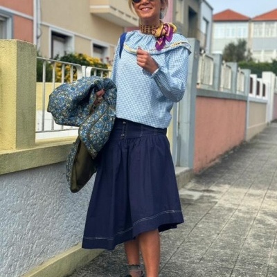 Mulher vestida com blusa azul claro, saia azul escura, lenço colorido no pescoço, sapatos castanhos, casaco azul floral, em rua pavimentada