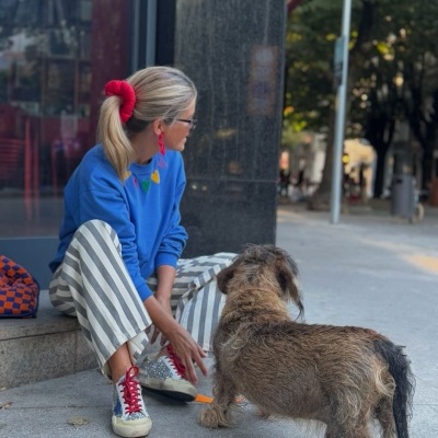 Mulher com camisola azul e calças riscas sentada na rua com cão de pelo castanho