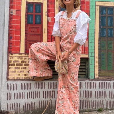 Mulher com macacão floral rosa, blusa branca, óculos de sol e bolsa, em frente a mural colorido