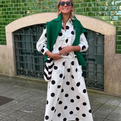 Mulher com roupa branca de bolinhas pretas, casaco verde e acessórios coloridos numa rua com azulejos verdes