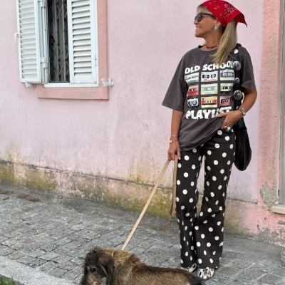 Mulher de lenço vermelho e óculos, t-shirt estampada e calças de bolinhas, com cão castanho em passeio junto a muro rosa com janela branca
