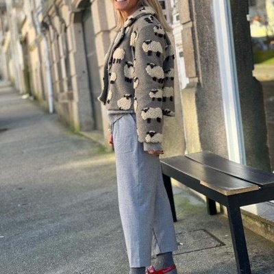 Mulher com casaco bege com padrão de ovelhas, calças cinza e sapatos vermelhos junto a um banco preto numa rua urbana