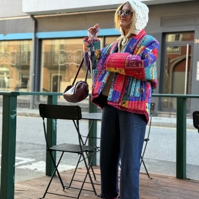Mulher com casaco colorido matelassado, calças azuis, chapéu branco de malha, mala castanha e mocassins castanhos