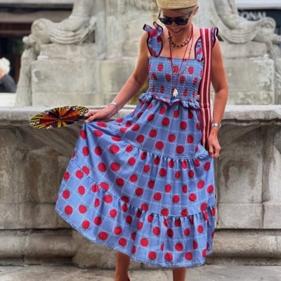 Mulher com vestido azul e bolas vermelhas, sapatos vermelhos e chapéu de palha, segurando leque colorido, junto a fonte em pedra.