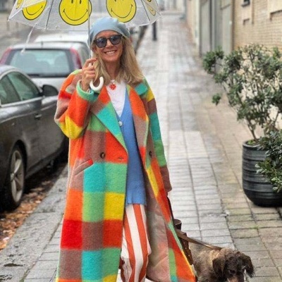 Mulher com casaco xadrez colorido e guarda-chuva transparente com sorrisos amarelos, passeando cão na rua molhada.