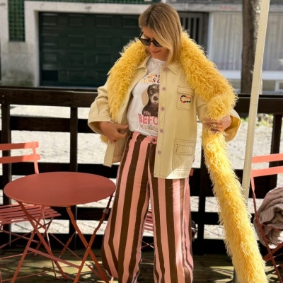 Pessoa com casaco bege e pelagem amarela, t-shirt com cão, calças às riscas rosa e castanho e sapatilhas ao lado de mesa vermelha ao ar livre