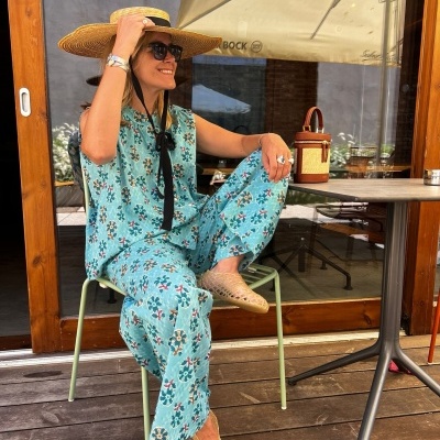 Mulher sentada de roupa azul floral, chapéu de palha e óculos escuros num espaço exterior com cadeira e mesa.