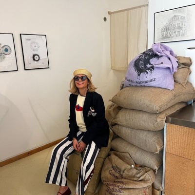 Mulher sentada ao lado de sacos de juta empilhados, com saco roxo estampado, calças de riscas verticais e blazer preto.