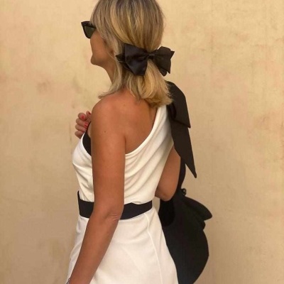 Mulher de cabelo louro com vestido branco de alça única, cinto preto, e fita preta no cabelo com laço