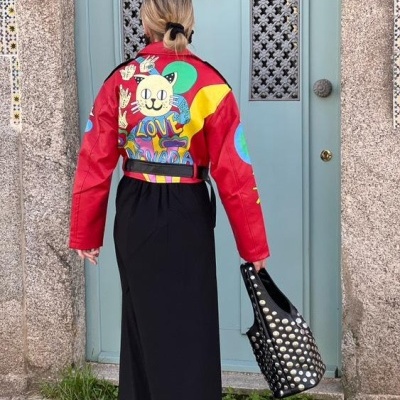 Mulher de costas com casaco colorido, vestido preto e carteira preta com tachas em frente a porta azul.