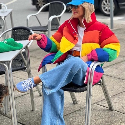 Mulher com casaco acolchoado colorido, boné azul e calças de ganga sentada numa cadeira ao ar livre