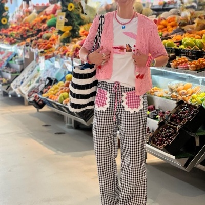 Mulher vestida com cardigan cor-de-rosa, t-shirt branca, calças xadrez e óculos escuros numa feira de fruta.