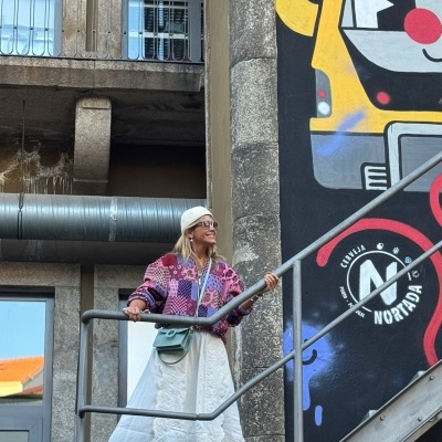 Mulher com vestido branco e casaco xadrez roxo numa escada metálica junto a mural graffiti colorido