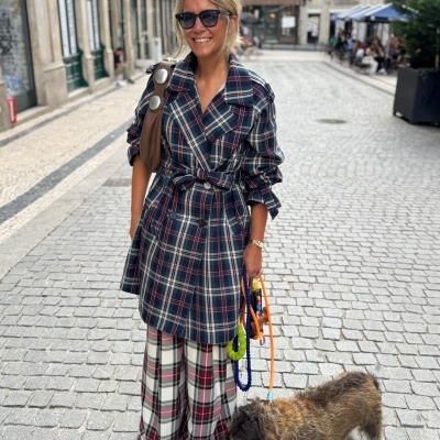 Mulher com casaco e calças xadrez, óculos escuros, mala castanha e cão com trelas coloridas numa rua de calçada portuguesa