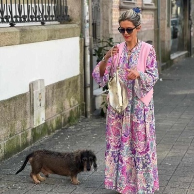 Mulher com vestido floral rosa e lilás, colete rosa, sapatos dourados e bolsa dourada, com cão pequeno numa rua de calçada