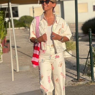 Mulher sorridente com conjunto estampado branco, bolsa rosa/vermelha de crochê e óculos escuros num local urbano