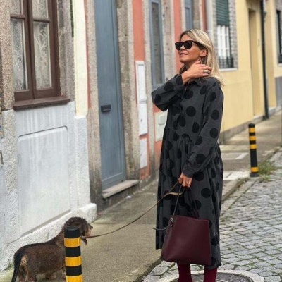 Mulher com vestido preto e bolinhas a passear com cão numa rua com calçada e paralelepípedos