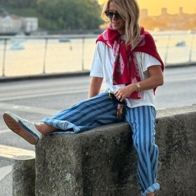 Mulher vestindo t-shirt branca, calças azuis riscas verticais, casaco vermelho ao ombro, óculos escuros, sentada em muro de pedra junto a rio com barcos e ponte ao fundo