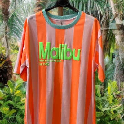 T-shirt listrada alaranjada e rosa claro com texto verde Malibu pendurada contra fundo de plantas tropicais