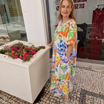 Mulher com vestido longo floral colorido ao lado de vaso branco com flores na calçada.