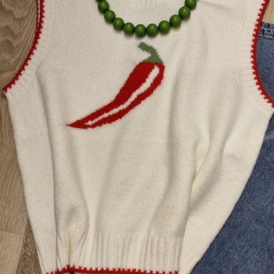 Camisola branca sem mangas com pimento bordado, colar verde e roupa azul junto, sobre chão de madeira.