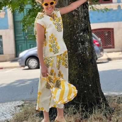 mulher com conjunto floral amarelo e branco, óculos amarelos, bolsa listrada amarela e branca junto a árvore na rua