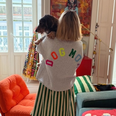 Mulher de costas com casaco cinzento e letras coloridas DOG MOM, saia verde e branca, segurando cão, em ambiente interno colorido.