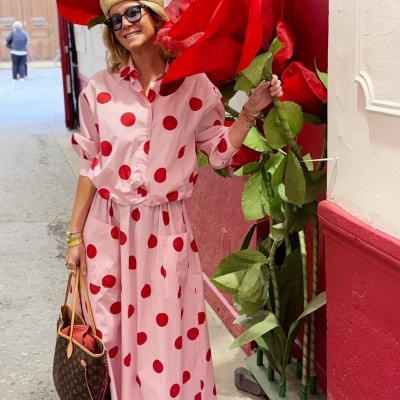 Mulher com vestido rosa de bolinhas vermelhas e chapéu de palha segurando flores vermelhas gigantes