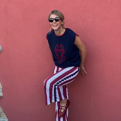 Mulher com colete azul e lagosta vermelha, calças listradas vermelhas e brancas, sapatos vermelhos, óculos de sol, encostada em parede cor de rosa