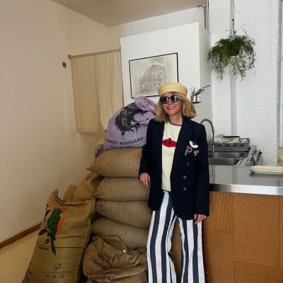 Mulher de casaco azul, chapéu de palha e calças listradas junto a sacos de juta e cozinha com pia de aço inox