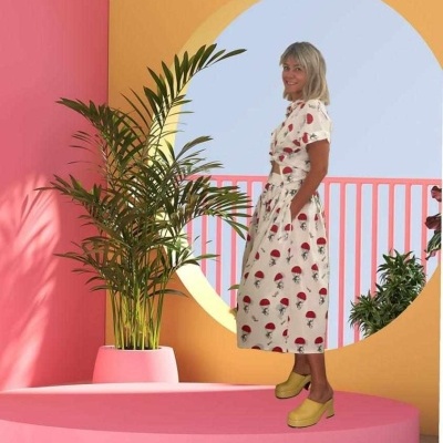 Vestido branco com padrão de cogumelos vermelhos e pretos e sapatos amarelos num cenário colorido com plantas e paredes coloridas.