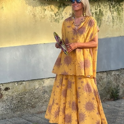 Mulher com roupa amarela estampada de sóis, óculos azuis, leque colorido e colares ao ar livre