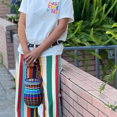 Pessoa vestindo t-shirt branca com texto colorido, calças com riscas coloridas e bolsa de palha colorida ao ar livre