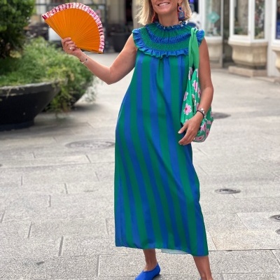 Mulher com vestido azul e verde, leque laranja, bolsa verde com flores e sapatos azuis numa rua pedonal.