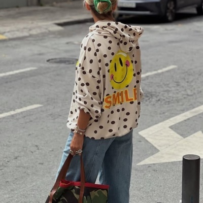 Pessoa vista de costas com casaco creme às bolinhas pretas, calças de ganga azuis e bolsa camuflada com carinha sorridente amarela