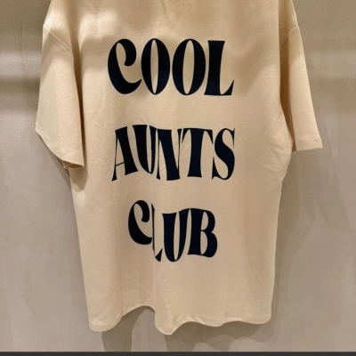 T-shirt oversize bege com texto preto nas costas pendurada na parede.