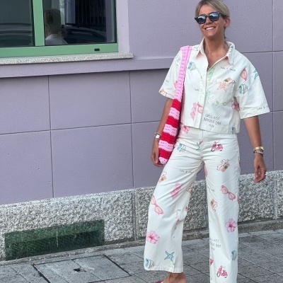 Mulher com conjunto branco estampado de camisa e calças largas, chinelos rosa e mala tricotada às riscas, encostada a parede roxa.