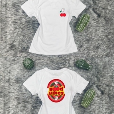 Camisola branca com design de cerejas e flores pequenas dianteira e good vibes em amarelo dentro de círculo vermelho na parte traseira, sobre superfície cinzenta e cercada por cactos verdes