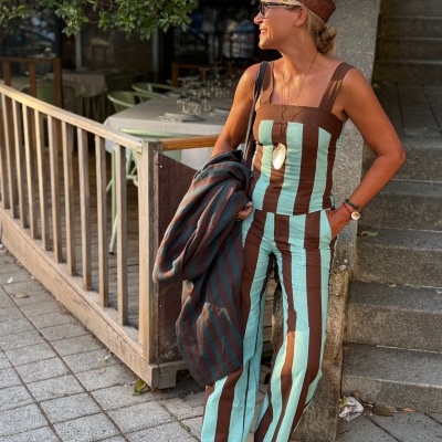 Mulher com conjunto de roupa riscada castanho e verde-água, chapéu castanho, óculos escuros pretos e sandálias pretas num cenário exterior de restaurante.