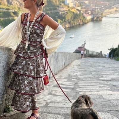 Mulher com vestido floral castanho e vermelho, lenço branco e colar, segurando trela de cão castanho numa estrada com vista para rio e cidade.